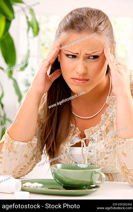 Young woman having headache, holding her head, leaning on table. Taking pills with tea