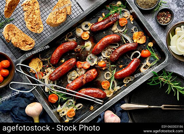 Top view on delicious grilled sausages cooked on barbecue served with onion, tomatoes, garlic, bread and herbs. Placed on metal tray
