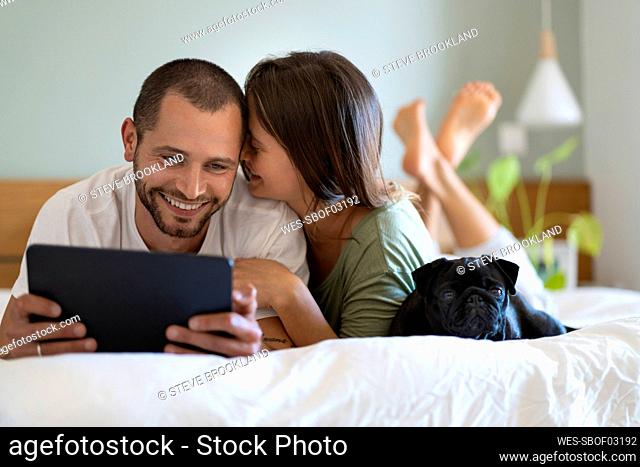 Woman embracing boyfriend looking at digital tablet while lying on bed in bedroom at home