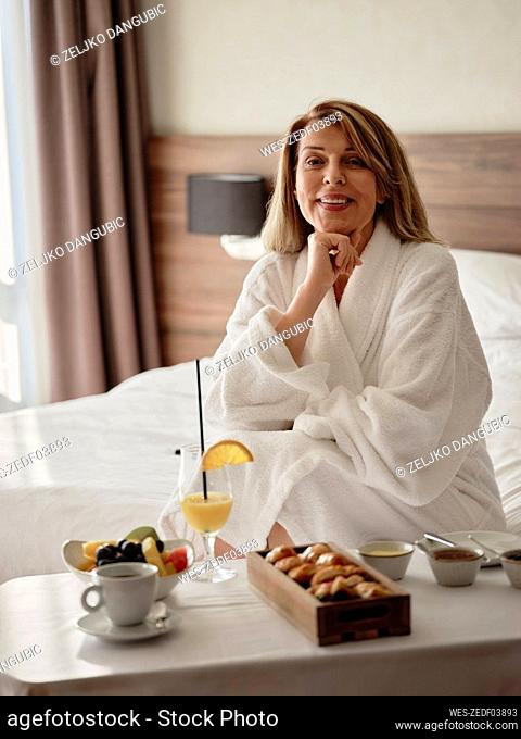 Smiling relaxed blond woman sitting with breakfast in hotel room