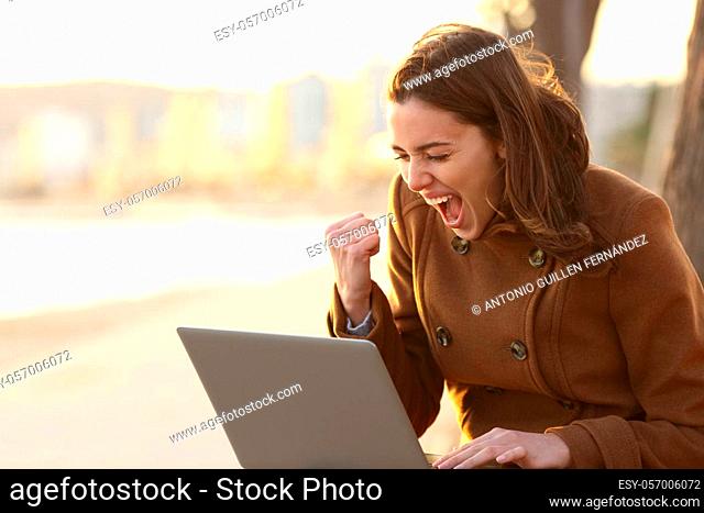 Excited woman reading good news on laptop sitting on a bench in winter on the beach