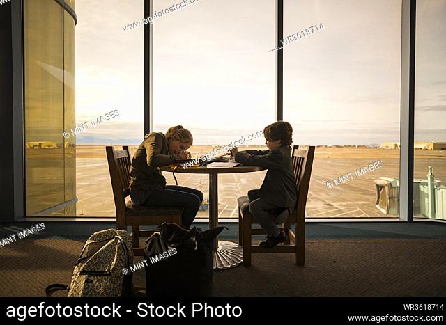 A boy and his older sister seated at a table in an airport lounge, writing and drawing