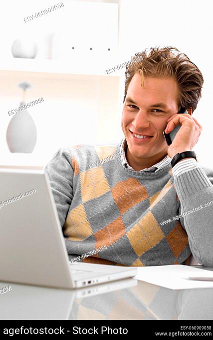Young man using laptop and calling on phone at home