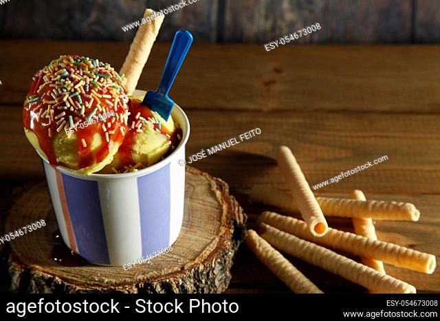 Two vanilla ice cream balls with sirope on ceramic bowl with blue plastic teaspoon and biscuit on wood slice and various biscuits on wooden background from...