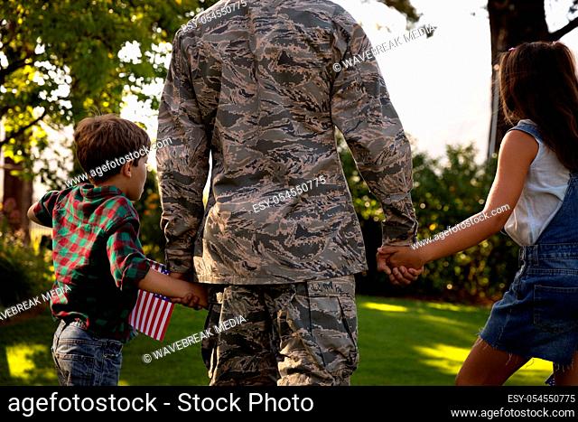 Rear view close up of a young adult mixed race male soldier in the garden outside his home, holding hands with his young son and daughter