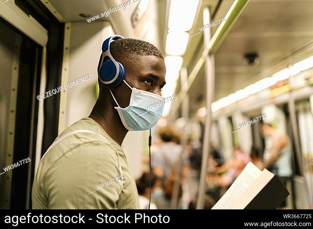 Close-up of young man wearing mask listening music through headphones looking away in train