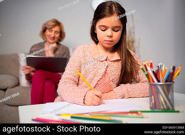 little girl drawing with pencils while her granny reading book on tablet behind