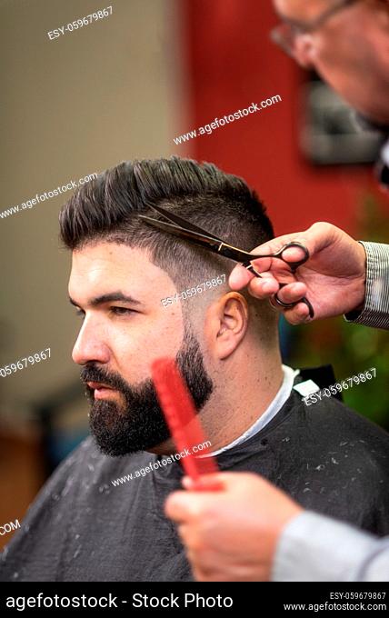 Handsome bearded man, having hair cut by scissors at barber shop