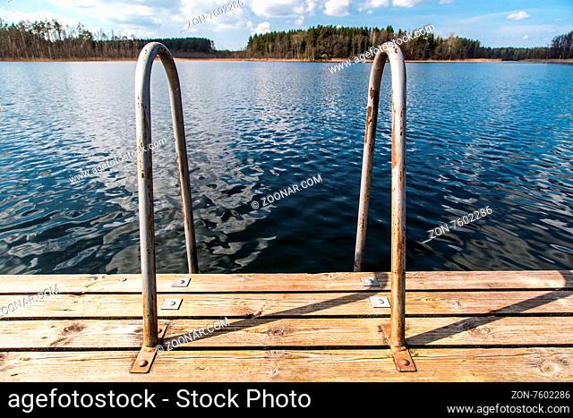 Close up detailed view of wooden pier with iron handrail inside lake, scenic view of reflection of the clouds and trees on lake, with cloudy blue sky background