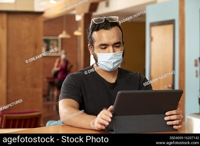 Young hispanic man wearing black t shirt and clear frame glasses with face mask, sitting at bar in kitchen of downtown loft with iPad