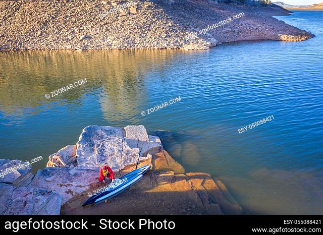 male paddler in a drysuit with hist stand up paddleboard on a rocky shore of mountain lake, aerial perspective