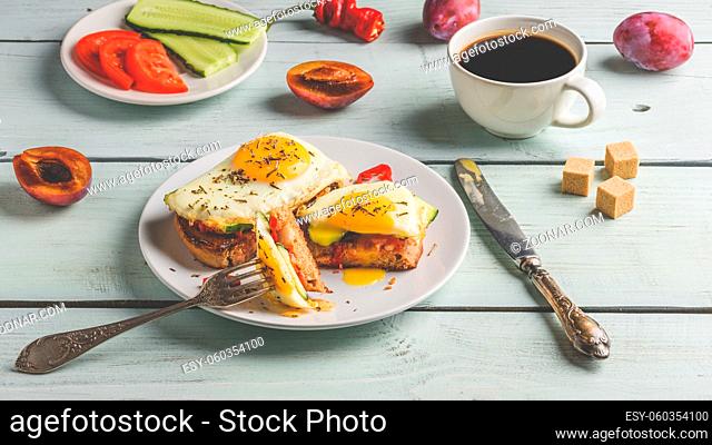 Sandwiches with vegetables and fried egg on white plate, cup of coffee and some fruits over wooden background