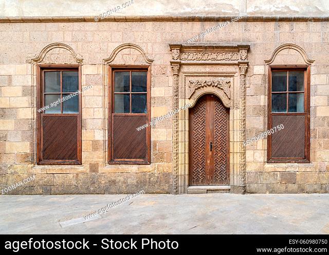 Facade of old abandoned stone decorated bricks wall with arched wooden door and three wooden shutter windows