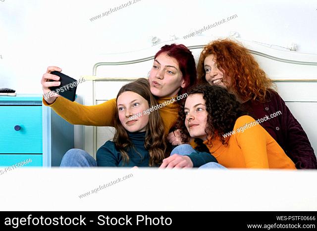 Mother and her three daughters sitting together on bed taking selfie with smartphone
