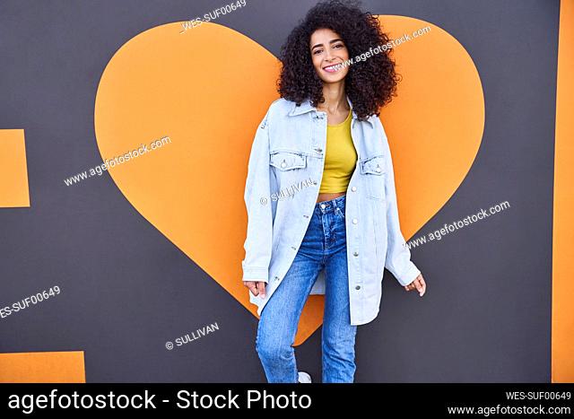 Happy young woman with curly hair standing against heart shape on wall