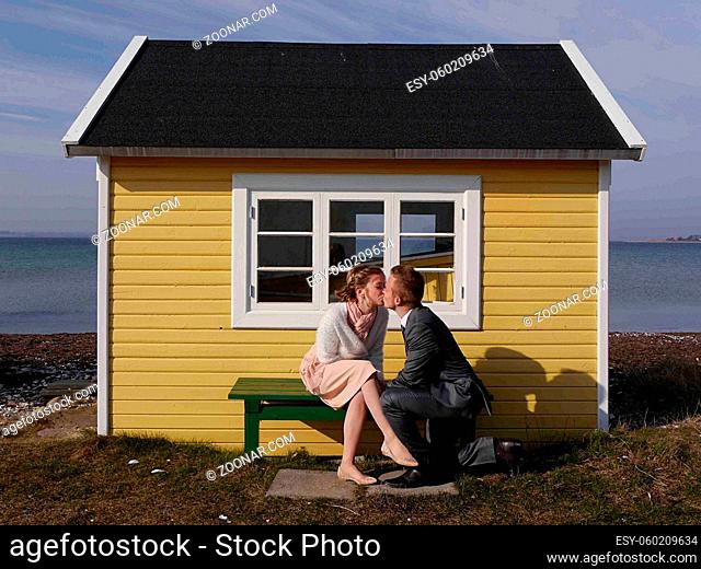 An attractive happy young husband on one knee kissing his beautiful girlfriend in front of cute, yellow beach hut for fun on Danish island of Aero