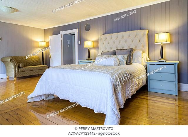 Queen size bed with white bedspread, beige cloth headboard, light blue end tables with drawers and gold and grey upholstered recamier in master bedroom with...