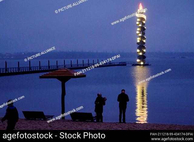 18 December 2021, Saxony-Anhalt, Muldestausee: Strollers on Lake Goitzsche with the water level tower in the background. The 26-meter-high structure floating on...