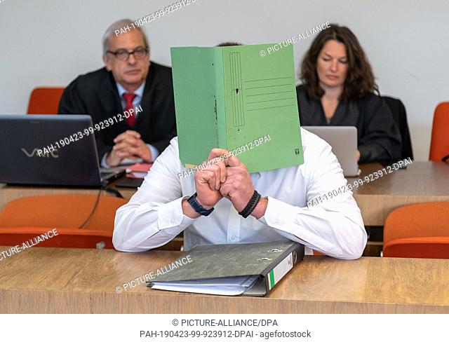 23 April 2019, Bavaria, München: The defendant holds a file cover in front of his face before the trial begins. Behind him are his lawyers Klaus Wittmann and...
