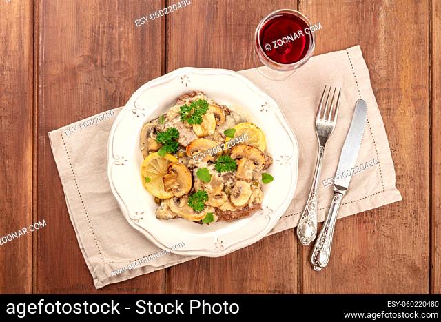 Scallopine di vitello, a traditional Italian dish of veal, with a mushroom sauce, lemons, and parsley, shot from above on a dark rustic wooden background with...