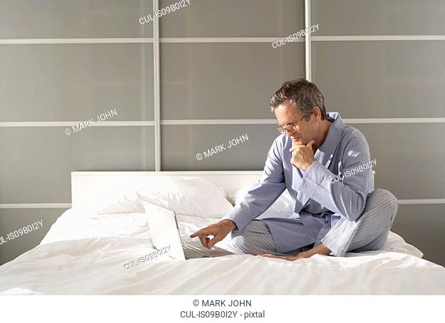 Puzzled senior man on bed pointing at laptop
