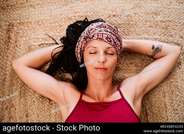 Woman wearing headscarf resting with hands behind head while lying on carpet at home