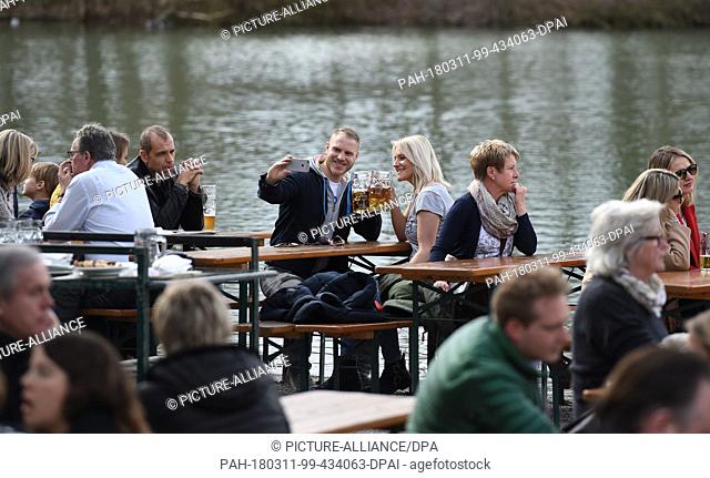 11 March 2018, Germany, Munich: A couple sitting in a beer garden at the Seehaus am Kleinhesseloher See in the Englischer Garten take a selfie