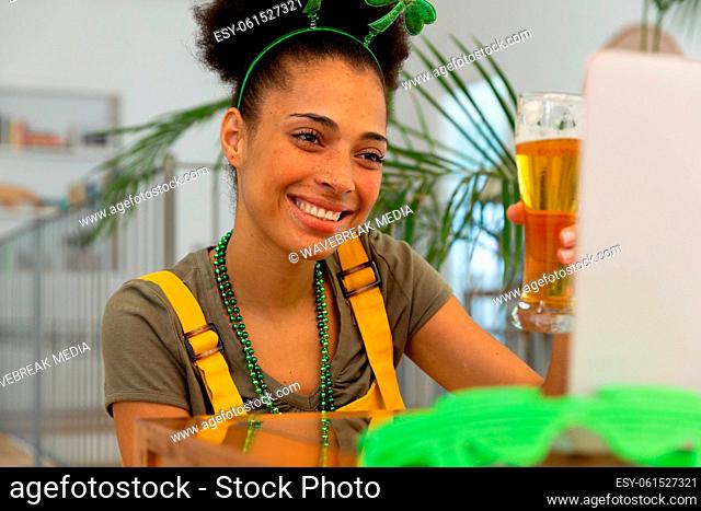 Happy mixed race woman celebrating st patrick's day making video call raising glass of beer