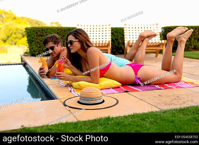 Side view of a Caucasian couple wearing beachwear and sunglasses in a garden, lying beside a swimming pool, holding cocktails and sunbathing