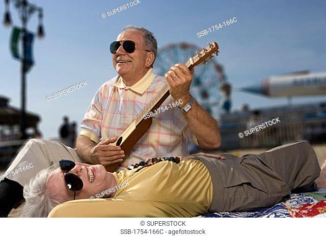 Senior man playing a guitar with a senior woman lying beside him, The Wonder Wheel, Coney Island, New York City, New York, USA