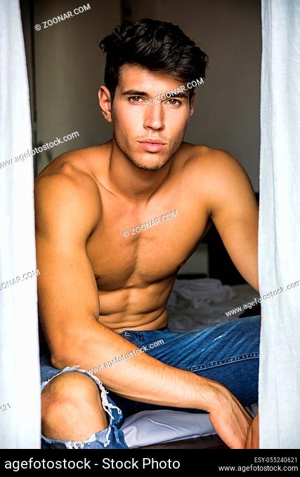 Sexy handsome young man sitting shirtless in his bedroom next to window curtains