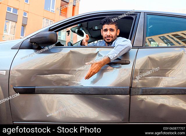 Portrait of confused handsome bearded man wearing blue shirt sitting behind the wheel of dented car with puzzled facial expression