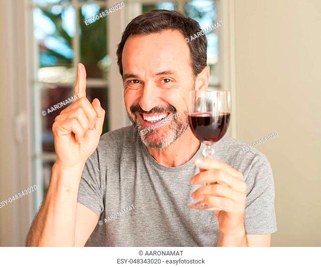 Middle age man drinking a glass of wine surprised with an idea or question pointing finger with happy face, number one