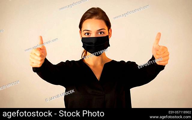 Positive woman wearing black face mask showing her thumbs up. Happy young Caucasian girl expressing encouragement and support with her fingers during medical...