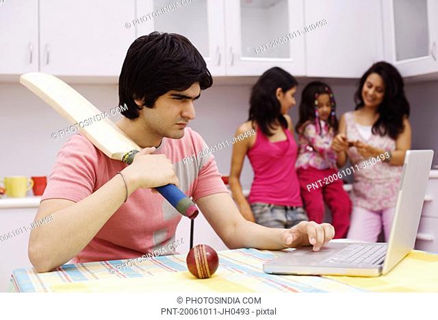 Young man holding a cricket bat and using a laptop in the kitchen