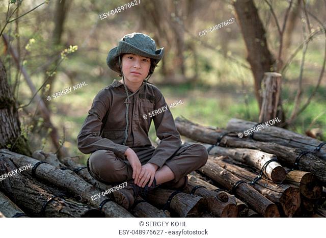 A boy in a coveralls and a hat sitting on a pile of sawn logs in the forest