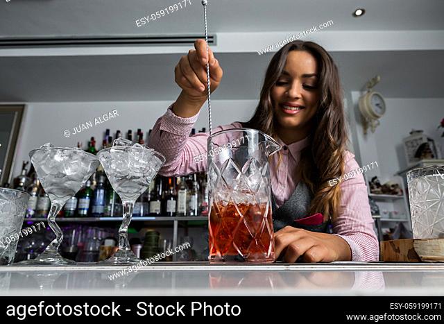 Low angle of cheerful young ethnic female bartender mixing alcoholic drinks in glass with long spoon while preparing cocktail at bar counter