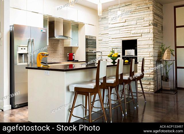Kitchen with white lacquered wooden cabinets, island with quartz countertop and high back barstools, recessed toaster and wine rack in insert of cut stone...