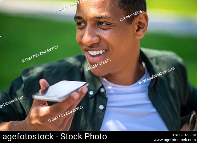 Wellbeing. Smiling dark-skinned guy recording voice message on smartphone looking away outdoors on sunny day