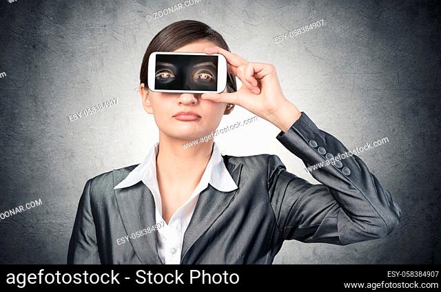 Portrait of pretty woman covering her eyes with smartphone. Businesswoman showing mobile phone with eyes on screen. Corporate businessperson on grey wall...