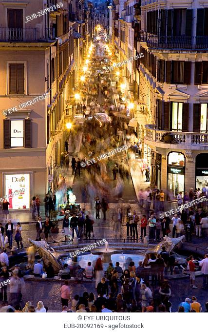 The Via dei Condotti the main shopping street busy with people illuminated at night seen from the Spanish Steps with seated tourists and the Fontana della...