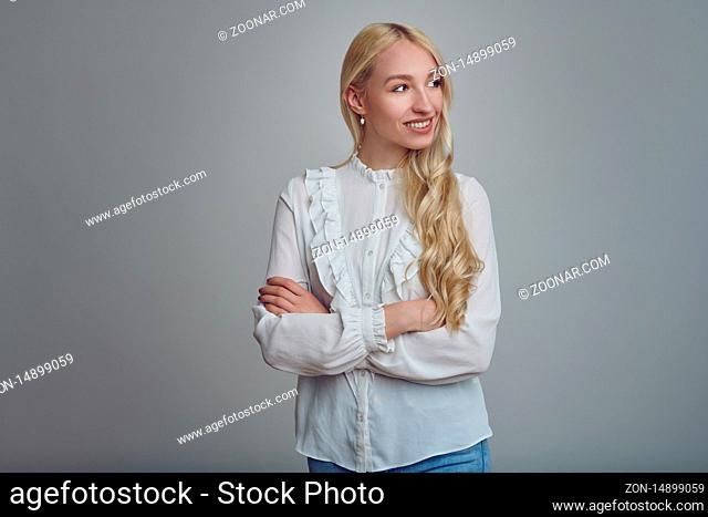 Young and beautiful long-haired blond woman in white blouse standing with her arms folded and looking to camera right with a smile