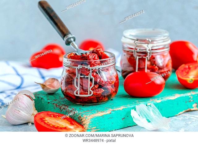 Fork in a jar of sun-dried tomatoes with spices and olive oil on an old wooden board, fresh tomatoes and a head of garlic on a grey concrete background