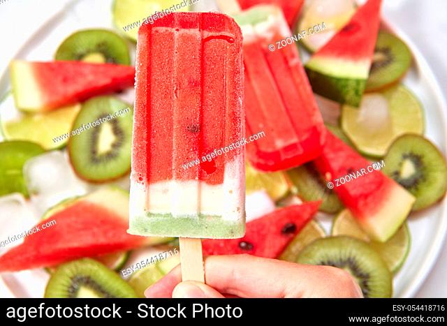 Close-up the girl's hand holds a homemade vitamin, frozen juice on a stick against the background of juicy organic slices of watermelon, lime