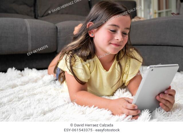 Cute little girl watch cartoons on digital tablet. Kid lies on floor laughing using electronic device. Indoor leisure for children