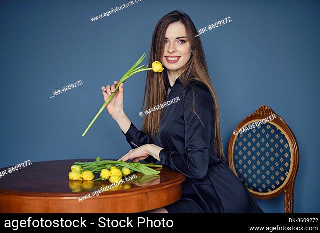 Beautiful young woman sits behind the table in victorian style with yellow tulips in hand
