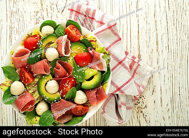 Healthy green salad with prosciutto, avocado and mozzarella cheese on wooden table background. Healthy meal