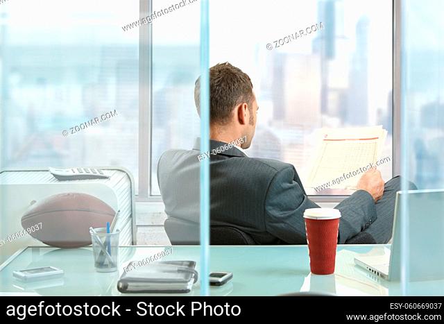 Relaxed businessman sitting at desk in front of office windows, reading nwespaper