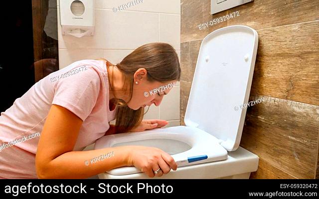 Young pregnant woman vomitting and suffering from nausea in toilet holding positive pregnancy test. Intoxication and nausea during pregnancy