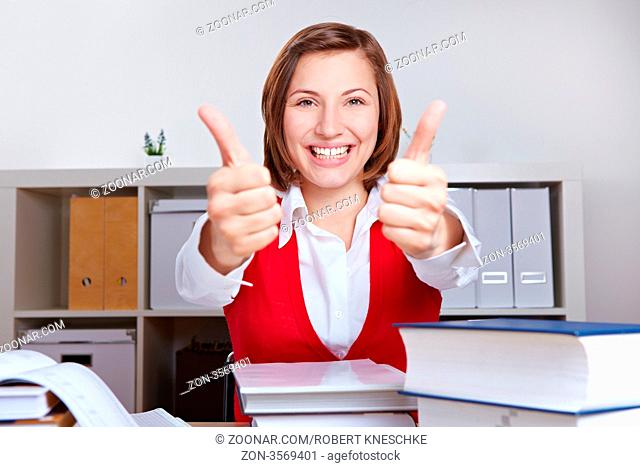 Lachende Frau beim Lernen am Schreibtisch hält beide Daumen hoch